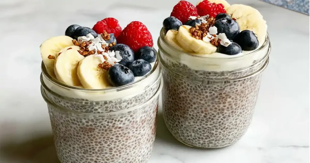 Whipped chia pudding in glass jars topped with banana slices, blueberries, raspberries, coconut flakes, and granola. Healthy no-bake breakfast or snack idea.